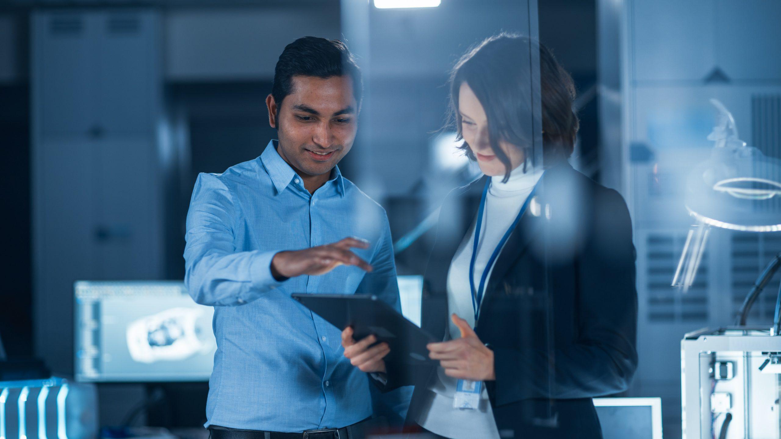 In Technology Research Facility: Female Project Manager Talks With Chief Engineer, they Consult Tablet Computer. Team of Industrial Engineers, Developers Work on Engine Design Using Computers